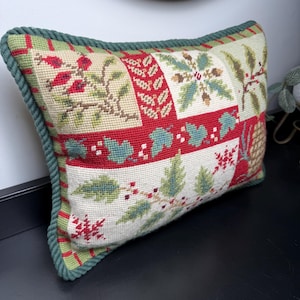 May include: A rectangular needlepoint pillow featuring a patchwork design of red, green, and cream botanical motifs including holly leaves, berries, and pinecones. The pillow is finished with a thick, dark green twisted rope trim border.