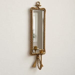 May include: A rectangular wall mirror with a gold-toned frame featuring a twisted rope design. The frame includes a decorative top loop and a bottom section with two candle holders and two hanging tassel-like ornaments in a matching gold finish.