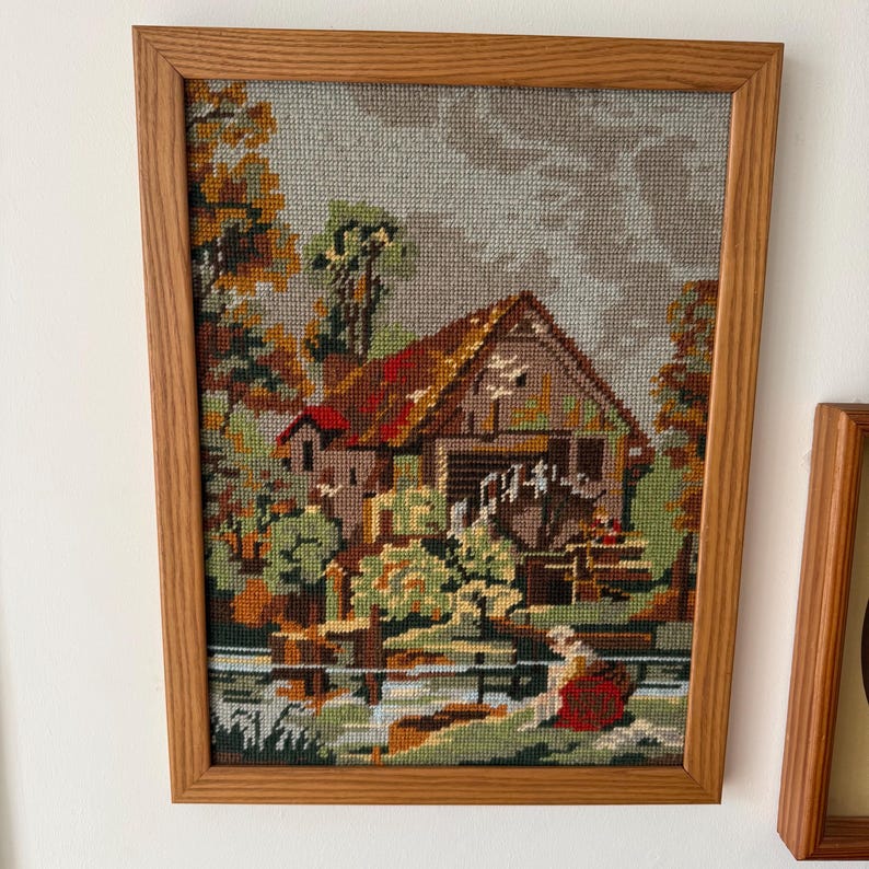 May include: Framed needlepoint art depicting a rural scene with a watermill, trees, and a figure by the water. The artwork features a brown wooden frame and a detailed, colorful landscape with a red-roofed building and lush greenery.