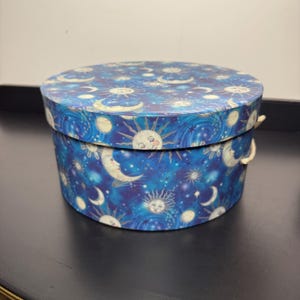May include: A round storage box with a lid featuring a celestial pattern of suns, moons, and stars against a deep blue background. The box has a cylindrical shape and includes a small, light-colored cord handle attached to the side of the base.