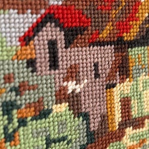 May include: Close-up of a cross-stitch design featuring a small house with a red roof and brown walls. The surrounding landscape includes green and brown foliage, all rendered in a textured, handmade style. The color palette is warm and earthy.