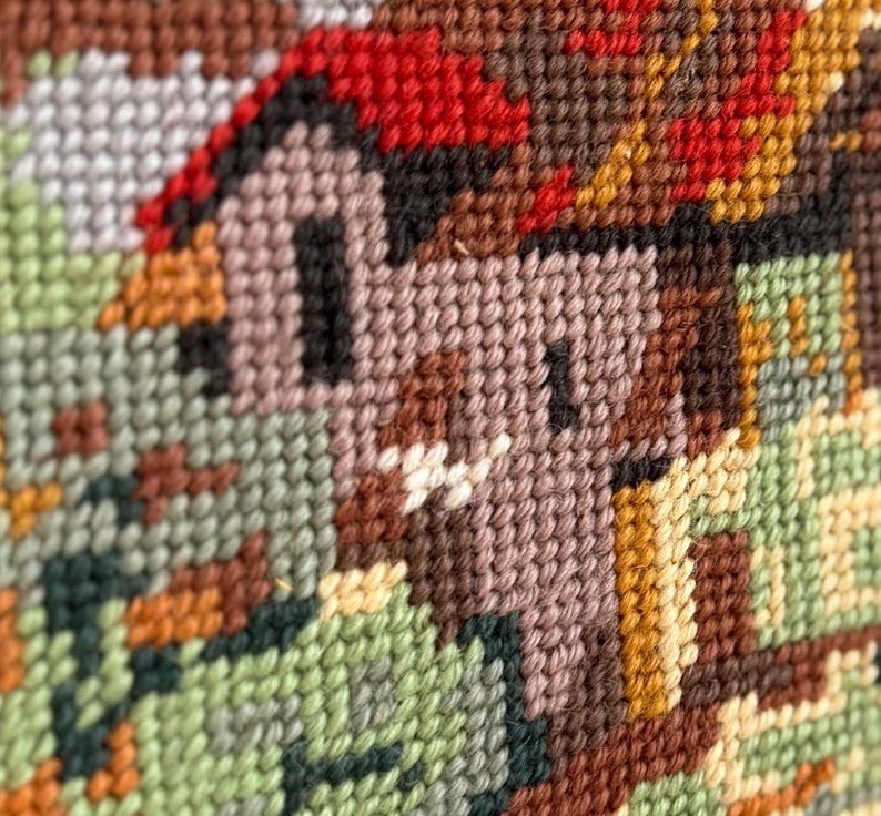 May include: Close-up of a cross-stitch design featuring a small house with a red roof and brown walls. The surrounding landscape includes green and brown foliage, all rendered in a textured, handmade style. The color palette is warm and earthy.