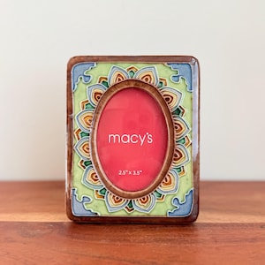 May include: A rectangular photo frame with a brown border and a decorative floral design in green, blue, and orange. The frame has an oval opening with the text "macy's" and dimensions of 2.5" x 3.5".