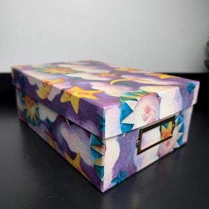 May include: A rectangular storage box with a lid featuring a purple and white cloudy sky pattern decorated with yellow stars and crescent moons. The front of the box includes a rectangular brass-colored metal label holder attached to the side.