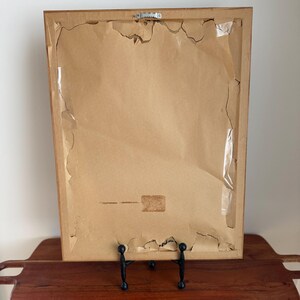 May include: Back of a framed artwork, showing the cardboard backing with torn edges and tape. The frame is a light brown color, and the artwork is displayed on a wooden tray with a black metal stand.