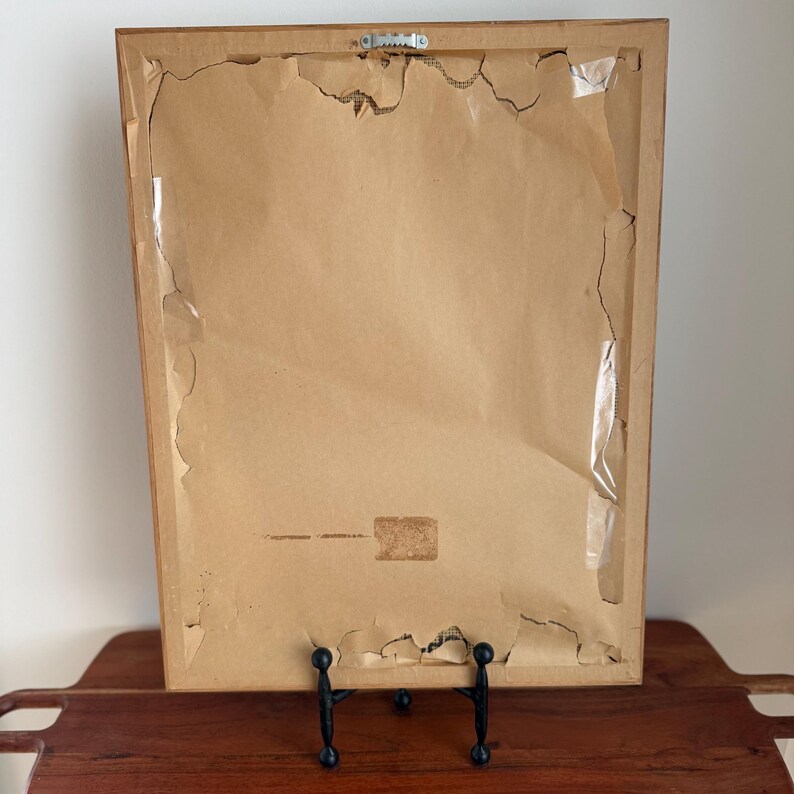 May include: Back of a framed artwork, showing the cardboard backing with torn edges and tape. The frame is a light brown color, and the artwork is displayed on a wooden tray with a black metal stand.