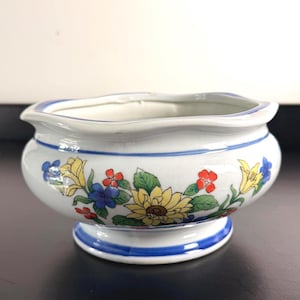 May include: A white ceramic bowl with a scalloped rim and a pedestal base. The exterior features a colorful floral pattern with yellow sunflowers, blue flowers, red blossoms, and green leaves, accented by thin blue horizontal bands around the rim and base.