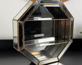 May include: An octagonal display case featuring mirrored glass panels framed in thin gold-toned metal. The structure includes a hinged door on the left side and a single horizontal shelf inside, creating two distinct storage sections for small items.