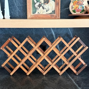 May include: A brown wooden accordion-style wine rack featuring a lattice design with multiple diamond-shaped storage slots. The rack is constructed from interconnected wooden slats joined by small metal rivets, creating an expandable structure.
