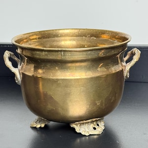 May include: A polished brass bowl with a rounded body, featuring two ornate side handles and three decorative feet. The metal surface displays a warm, golden tone with subtle variations in texture and finish across the exterior and rim.