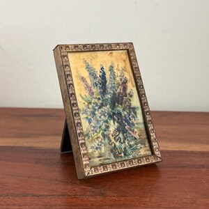 May include: A small framed painting of blue and purple flowers in a vase. The painting is in a rectangular frame with a decorative border. The frame is supported by a small easel-style stand. The painting is on a wooden surface.