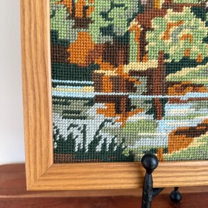 May include: A framed needlepoint landscape featuring a river scene with trees. The artwork uses various shades of green, brown, and orange, with a light blue and white river. The frame is made of light-colored wood.