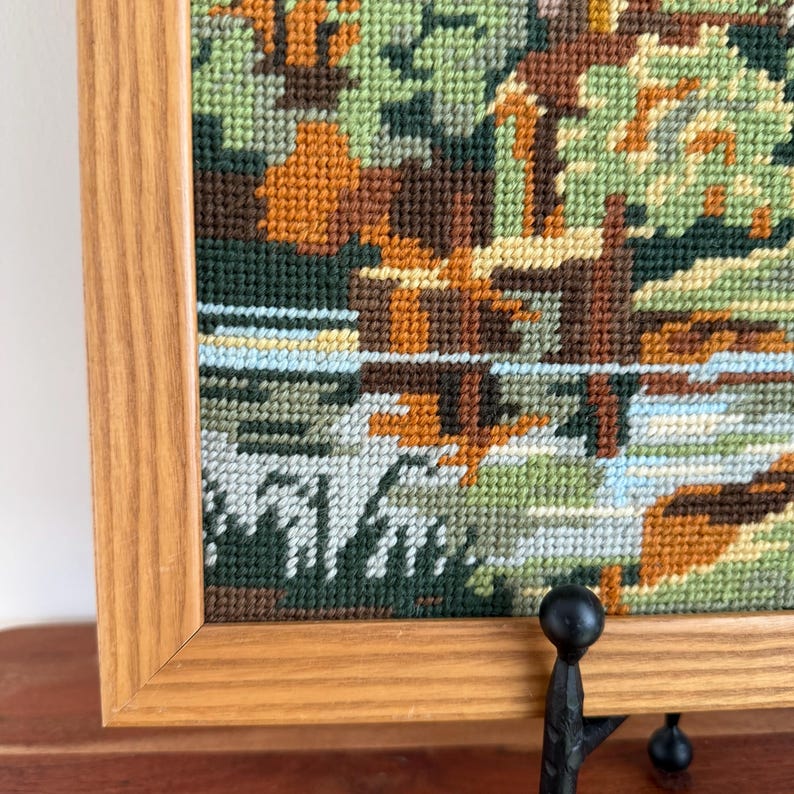 May include: A framed needlepoint landscape featuring a river scene with trees. The artwork uses various shades of green, brown, and orange, with a light blue and white river. The frame is made of light-colored wood.