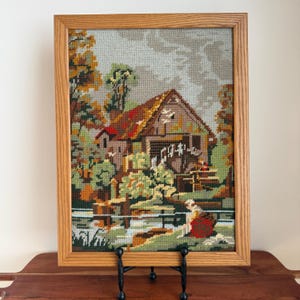 May include: A framed needlepoint scene of a water mill in a rural landscape. The artwork features a wooden mill with a red roof, surrounded by trees in shades of green, orange, and brown. The frame is made of light-colored wood.