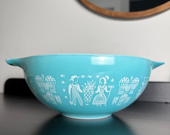 May include: A turquoise mixing bowl with two side handles, featuring a white screen-printed pattern of a farmer and a person holding a basket of produce, surrounded by wheat sheaves and small garden tools, set against a solid turquoise background.