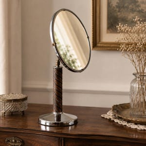 May include: A round tabletop mirror with a silver-toned metal frame and a circular base. The central support column features a dark brown, spiraled wooden design. The mirror is adjustable, mounted on a swivel mechanism that allows for tilting.