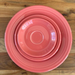 May include: Overhead view of a stack of coral-colored ceramic dishware on a wooden surface. The set includes a large plate, a smaller plate, and a bowl. The dishes have a glossy finish and a ridged design around the edges.