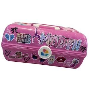 May include: A pink, rectangular storage case with a white handle and a white clasp. The case is decorated with stickers, including the name "MADYN" in glittery letters, hearts, and the words "CAMP VIBES".
