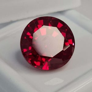 Peut inclure: Une pierre précieuse ronde à facettes d'une couleur rouge profond et vibrante. La pierre présente une taille brillant avec de multiples facettes triangulaires et trapézoïdales qui reflètent la lumière, créant un contraste de tons rouges.