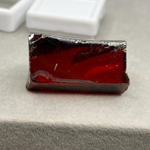 May include: A rectangular piece of deep red, translucent material with a smooth, glossy surface and slightly irregular, sharp edges. The object displays variations in colour intensity, ranging from dark crimson to lighter ruby tones throughout.