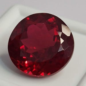 May include: A round, faceted gemstone with a deep, vibrant red colour. The stone features a brilliant cut with numerous triangular and kite-shaped facets that reflect light, creating a sparkling appearance across its polished, translucent surface.