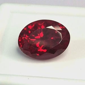 Può includere: Una gemma sfaccettata, di forma ovale, di colore rosso intenso. La gemma ha una superficie lucida con molteplici sfaccettature che riflettono la luce, creando un effetto scintillante. La pietra è su uno sfondo bianco.