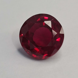Könnte beinhalten: Ein runder, facettierter Edelstein in einer tiefen, gesättigten roten Farbe. Der Stein weist einen Brillantschliff mit mehreren dreieckigen und trapezförmigen Facetten auf, die das Licht reflektieren und ein klares, transparentes Aussehen erzeugen.