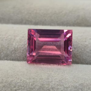 May include: A rectangular, faceted gemstone with a vibrant pink hue. The stone features a clear, transparent quality with sharp, geometric edges and a polished surface that reflects light, showcasing a deep, saturated magenta colour throughout.