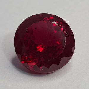 Könnte beinhalten: Ein runder, facettierter Edelstein in einer tiefen, gesättigten roten Farbe. Der Stein weist einen Brillantschliff mit zahlreichen dreieckigen und rautenförmigen Facetten auf, die das Licht reflektieren und ein komplexes Muster erzeugen.