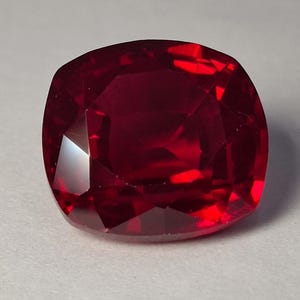 Può includere: Una pietra preziosa con taglio a cuscino di colore rosso intenso e saturo. La pietra presenta una superficie sfaccettata che riflette la luce, creando distinti motivi geometrici e riflessi sulla sua struttura trasparente, lucida e simmetrica.