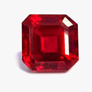 以下が含まれることがあります： 正方形にカットされた、深い赤色の宝石。宝石は、面取りされたエッジと中央の正方形のファセットを持つ長方形の形状をしています。石は非常に反射性が高く、内部で光が反射し、白い背景に配置されています。