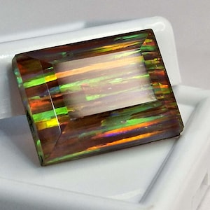 May include: A rectangular faceted gemstone with a dark base color and vibrant flashes of green, orange, and yellow light. The stone features a polished surface with sharp edges and a clear play of color across its entire top surface area.