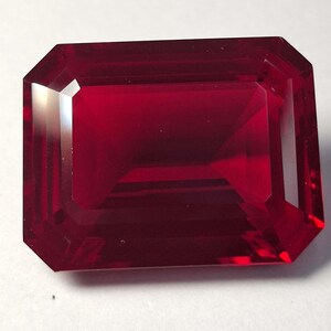 May include: A rectangular, emerald-cut gemstone with a deep, vibrant red colour. The stone features precise, multi-faceted edges that create a clear, reflective surface with internal light refraction visible throughout the transparent, polished body.