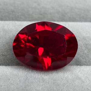 May include: A vibrant, deep red gemstone cut into a smooth oval shape with multiple facets that reflect light. The stone displays a rich, saturated crimson hue throughout its surface, showcasing a clear, transparent quality with sharp geometric edges.