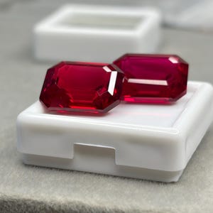 Peut inclure: Deux pierres précieuses rouge vif, taillées en émeraude, présentant des facettes nettes et une finition brillante. Ces pierres rectangulaires sont posées sur un petit récipient de rangement carré en plastique blanc avec un couvercle transparent.