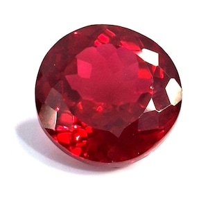 May include: A faceted, round-cut ruby gemstone. The gem is a deep, saturated red color, with multiple facets that reflect light. The ruby is set against a white background, highlighting its brilliance and clarity. This gemstone is suitable for jewelry.