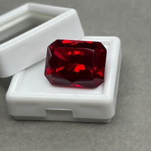 May include: A rectangular, faceted, deep red gemstone displayed inside an open white plastic storage box. The stone features sharp, geometric cuts that reflect light, creating a vibrant crimson appearance against the white container base.