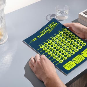 Könnte beinhalten: Eine Person hält einen blau-gelben 5K-Trainingsplan mit dem Titel "Zero To Hero". Der Plan enthält einen Wochenplan mit Lauf- und Ruhetagsindikatoren. Ein Glas Wasser und ein dekoratives Glas befinden sich im Hintergrund.