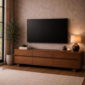 Könnte beinhalten: Modernes Wohnzimmer mit einem großen Fernseher über einem Holz-TV-Schrank mit sechs Schubladen. Ein beiger Teppich bedeckt den Boden, und eine Topfpflanze und eine Lampe ergänzen die Einrichtung. Der Schrank ist etwa 180 cm lang.
