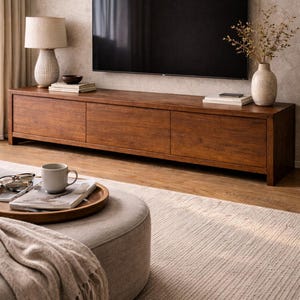 May include: A modern living room setup featuring a large, dark wooden TV console with three drawers. A flat-screen TV is mounted above. A beige rug, ottoman, and decorative vase with dried plants complete the scene. A lamp and books are on the console.
