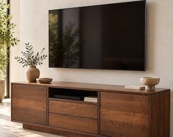 Könnte beinhalten: Ein langes, rechteckiges Medienboard aus dunklem Holz mit zwei Seitenschränken, zwei mittigen Schubladen und einem offenen Fach. Das Möbelstück steht unter einem großen Flachbildfernseher an einer hellen Wand.