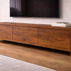 K&ouml;nnte beinhalten: Ein langes, rechteckiges Medienboard aus mittelbraunem Holz mit sichtbarer Maserung. Das M&ouml;belst&uuml;ck verf&uuml;gt &uuml;ber drei flache Schrankt&uuml;ren und steht auf kurzen, stabilen Beinen. Auf der rechten Seite der Oberseite liegt ein Stapel B&uuml;cher.