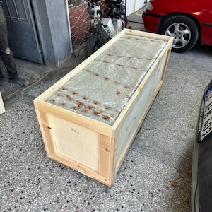 K&ouml;nnte beinhalten: Eine rechteckige Versandkiste aus hellem Naturholz, die einen gro&szlig;en, in Klarsichtfolie eingewickelten Gegenstand mit orangefarbenem Klebeband enth&auml;lt. Die Kiste besteht aus stabilen Holzbrettern und Holzplatten.