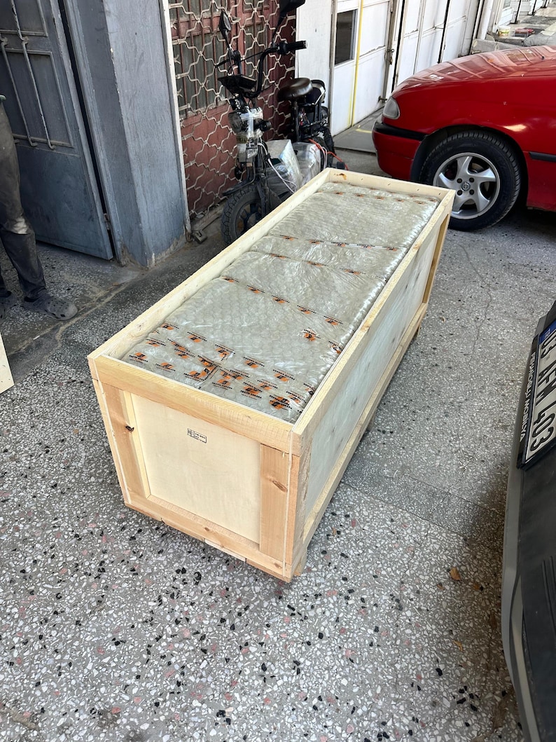 K&ouml;nnte beinhalten: Eine rechteckige Versandkiste aus hellem Naturholz, die einen gro&szlig;en, in Klarsichtfolie eingewickelten Gegenstand mit orangefarbenem Klebeband enth&auml;lt. Die Kiste besteht aus stabilen Holzbrettern und Holzplatten.