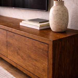 K&ouml;nnte beinhalten: Ein Medienkonsole aus dunklem Holz mit glatter Oberfl&auml;che und zwei Schrankt&uuml;ren. Darauf steht eine cremefarbene Keramikvase mit rissiger Textur, die getrocknete Zweige enth&auml;lt, sowie ein Stapel von drei B&uuml;chern mit neutralen Einb&auml;nden.