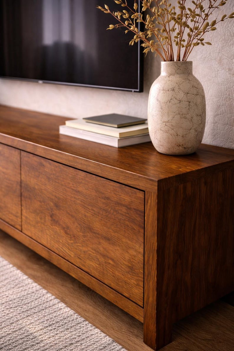 K&ouml;nnte beinhalten: Ein Medienkonsole aus dunklem Holz mit glatter Oberfl&auml;che und zwei Schrankt&uuml;ren. Darauf steht eine cremefarbene Keramikvase mit rissiger Textur, die getrocknete Zweige enth&auml;lt, sowie ein Stapel von drei B&uuml;chern mit neutralen Einb&auml;nden.