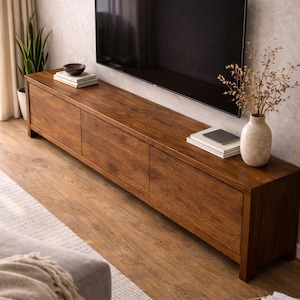 K&ouml;nnte beinhalten: Ein langes, rechteckiges Medienboard aus Holz mit warmbrauner Oberfl&auml;che und drei Schrankt&uuml;ren. Die Oberseite des Boards bietet Platz f&uuml;r eine kleine dunkle Schale auf einem B&uuml;cherstapel sowie eine helle Vase mit getrockneten Zweigen.