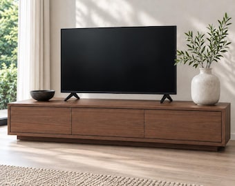 Könnte beinhalten: Ein niedriges, rechteckiges Medienmöbel aus Holz mit drei horizontalen Schubladen in dunkler Walnussoptik. Auf dem Möbel steht ein Flachbildfernseher, ergänzt durch eine kleine dunkle Schale und eine strukturierte weiße Vase mit Grün.