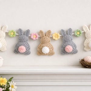 Peut inclure: Une guirlande de Pâques faite à la main avec des lapins crochetés blancs, gris et beiges, chacun avec une queue touffue. La guirlande est ornée de fleurs crochetées roses, jaunes et vertes. La guirlande est exposée au-dessus d'une étagère blanche avec des œufs de Pâques.