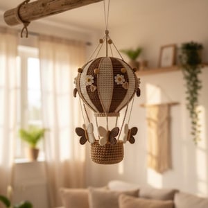 Op de afbeelding: Een handgemaakte gehaakte heteluchtballon mobiel in bruin en crème, versierd met madeliefjes en vlinderhangers. De mobiel hangt aan een houten balk, ideaal als speelse decoratie voor de kinderkamer.